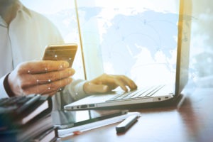 Person working at desk with cellphone in one hand and a laptop on the desk.